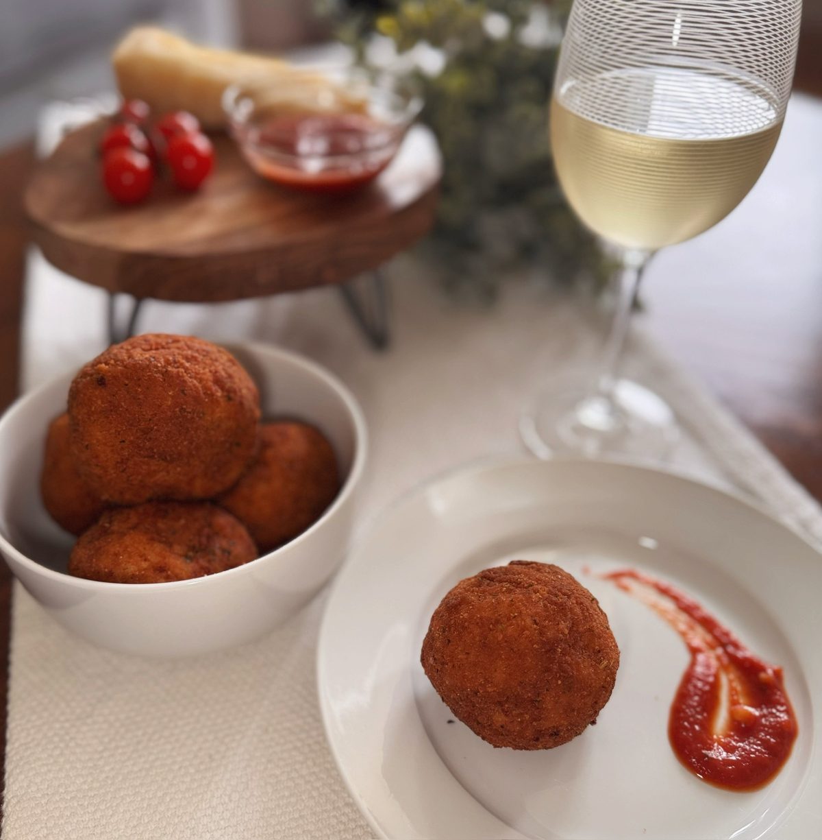 A plated arancini with marinara next to a bowl of arancini and a glass of wine
